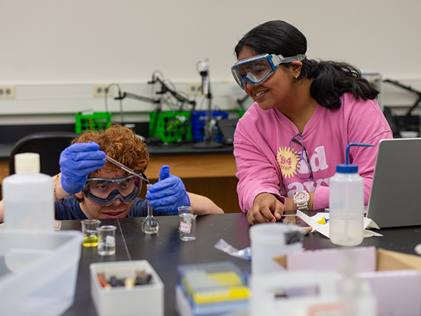 A female and male student interact in a Chemistry lab setting. Beakers and scientific instruments are arrayed on the table in front of them.